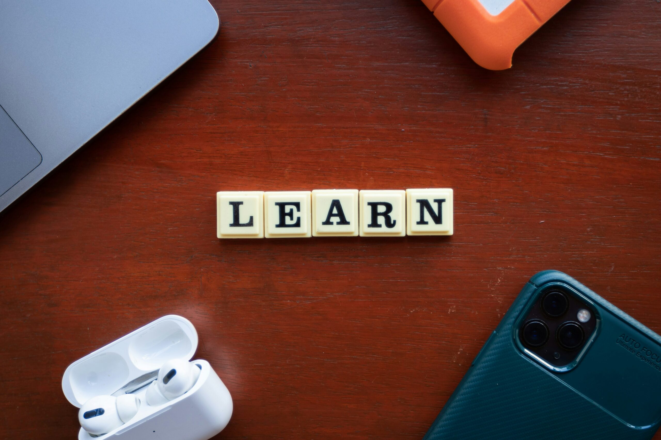 An image of the word LEARN made of block letter tiles laying on a mahogany wood desk. various electronics are distributed in the corners. a phone, earbuds, a laptop, and a tablet